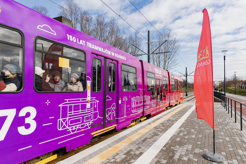 Na zdjęciu widzimy tramwaj ozdobiony fioletową grafiką upamiętniającą 150 lat tramwajów w Gdańsku, stojący na nowoczesnym peronie. Obok znajduje się czerwona flaga z herbem Gdańska, a w tle widoczne są gołe drzewa i otwarta przestrzeń.