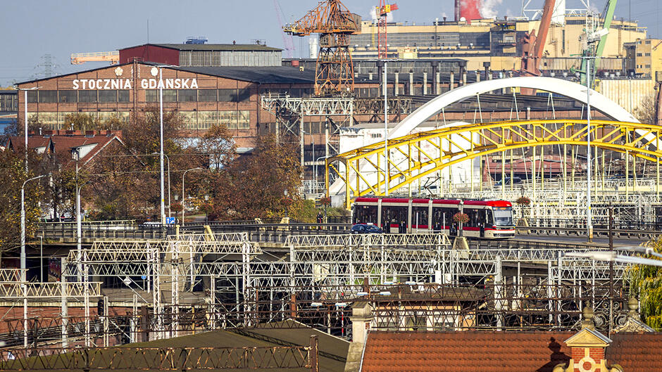 Zdjęcie przedstawia industrialny krajobraz stoczni w Gdańsku, w Polsce, z charakterystycznym napisem „Stocznia Gdańska” na fasadzie ceglanego budynku. W tle widoczne są dźwigi portowe, hale przemysłowe i inne elementy związane z działalnością stoczniową. Na pierwszym planie znajduje się żółty, łukowy most z białymi elementami konstrukcyjnymi, przez który przejeżdża czerwono-biały tramwaj. Wokół mostu znajdują się liczne metalowe konstrukcje i instalacje. Scena ma miejski, przemysłowy charakter z elementami infrastruktury transportowej i architektury historycznej. W tle widać dym unoszący się z kominów, co podkreśla funkcję przemysłową tego miejsca.