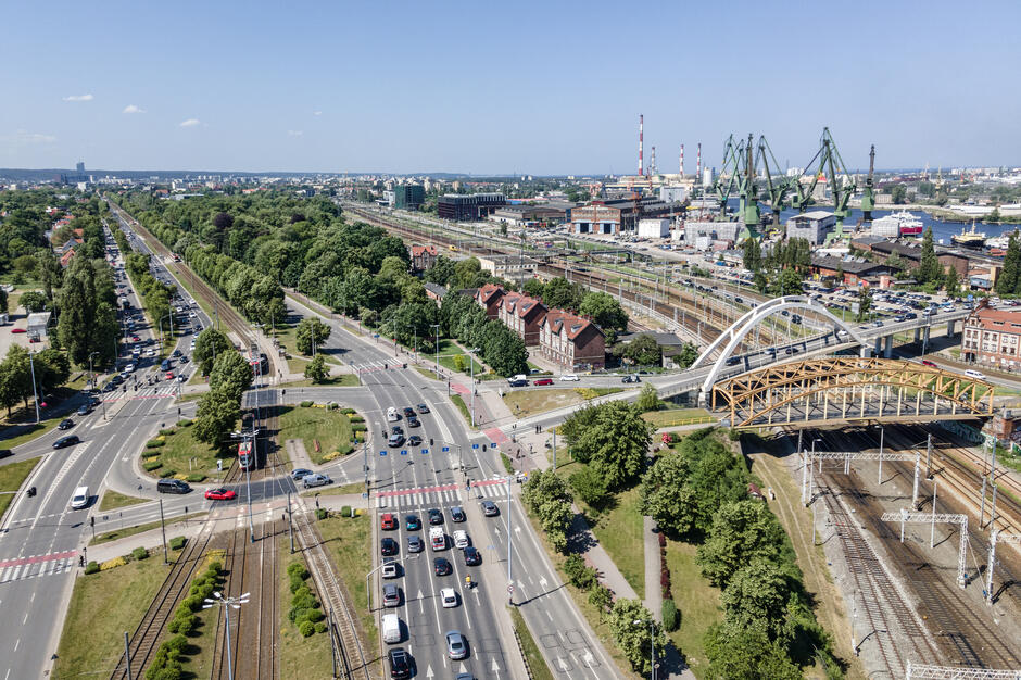 Zdjęcie wykonane z lotu ptaka ukazuje fragment Gdańska, prezentując miejski krajobraz z infrastrukturą drogową, kolejową oraz przemysłową. W dolnej części zdjęcia widoczny jest ruchliwy węzeł komunikacyjny z wieloma pasami ruchu, torowiskiem tramwajowym oraz rondem. W centralnej części widoczny jest biały i żółty most, który przecina linię kolejową i prowadzi w kierunku obszarów przemysłowych. Po lewej stronie kadru rozciągają się zielone tereny z alejami drzew, parkiem oraz linią tramwajową. Po prawej stronie widoczne są tory kolejowe, zabudowania mieszkalne z czerwonymi dachami oraz rozległy teren Stoczni Gdańskiej z charakterystycznymi żurawiami stoczniowymi i kominami przemysłowymi. Na horyzoncie rozpościera się panorama miasta z wieloma budynkami mieszkalnymi i biurowymi. Całość ukazuje harmonię między zielenią, infrastrukturą transportową i przemysłowym dziedzictwem miasta.