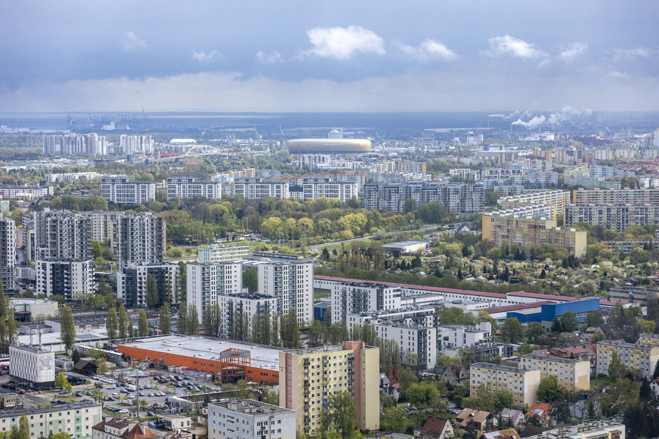 Zdjęcie przedstawia panoramę miejską Gdańska, ukazującą gęstą zabudowę mieszkaniową i przemysłową. Na pierwszym planie widoczne są bloki mieszkalne o różnej wysokości, w tym zarówno starsze, wielkopłytowe budynki, jak i nowoczesne apartamentowce. Pomiędzy budynkami znajdują się liczne tereny zielone, drzewa oraz niewielkie osiedla domków jednorodzinnych. W centrum kadru uwagę przyciąga Stadion Polsat Plus Arena Gdańsk, o charakterystycznym, bursztynowym kolorze, znajdujący się w dalszej części miasta. W tle, na horyzoncie, widać port morski z dźwigami i obiektami przemysłowymi, a także linię brzegową. Nad miastem unoszą się chmury, dodając scenerii dramatycznego klimatu, a w oddali widoczny jest dym z kominów przemysłowych. Całość oddaje kontrast między miejską infrastrukturą, naturą i przemysłowym charakterem regionu.