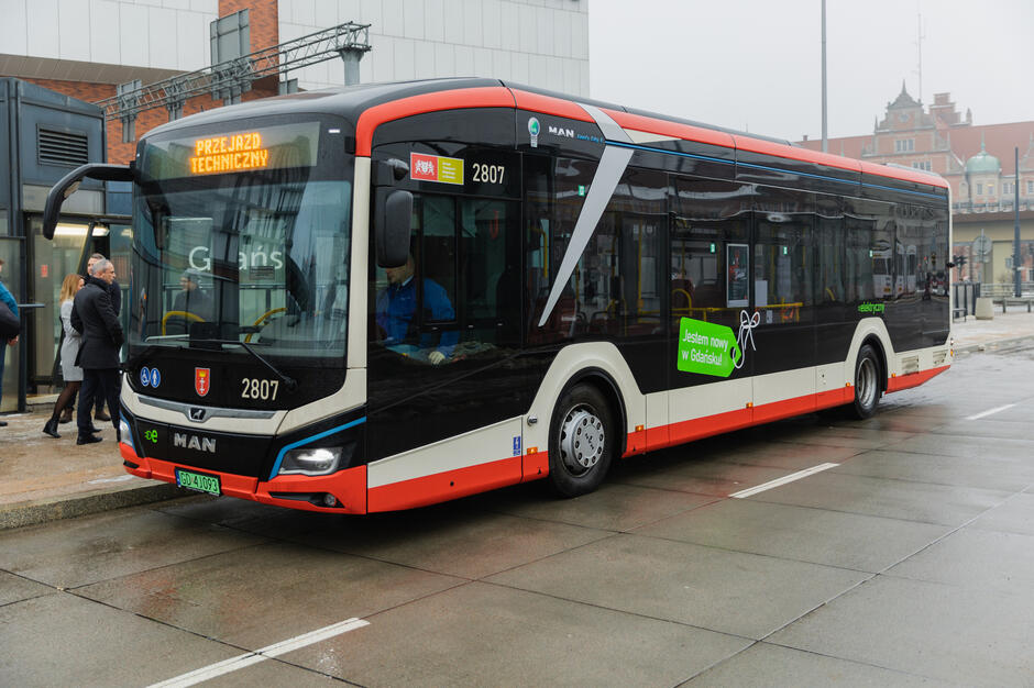 Zdjęcie przedstawia nowoczesny autobus marki MAN należący do komunikacji miejskiej w Gdańsku. Pojazd, oznaczony numerem bocznym 2807, jest elektryczny, co podkreśla napis elektryczny  widoczny na jego boku. Na wyświetlaczu z przodu autobusu widnieje komunikat „Przejazd techniczny”, co wskazuje, że autobus nie realizuje kursu z pasażerami. Na boku pojazdu znajduje się naklejka z napisem „Jestem nowy w Gdańsku!”, co sugeruje, że autobus dopiero został wprowadzony do floty.  Autobus ma nowoczesny design w biało-czarno-czerwonych barwach, charakterystycznych dla gdańskiej komunikacji miejskiej. W tle widoczny jest fragment miejskiego otoczenia, w tym przechodnie oraz budynki. Scena ma miejsce w pochmurny dzień, co widać po wilgotnej nawierzchni i zamglonym tle.       