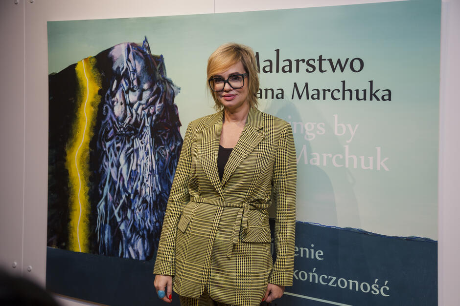 Na zdjęciu widać kobietę pozującą na tle plakatu promującego malarstwo Ivana Marchuka. Kobieta ubrana w elegancki żółto-czarny garnitur z wzorem w kratę stoi obok obrazu przedstawiającego abstrakcyjną, dynamiczną kompozycję