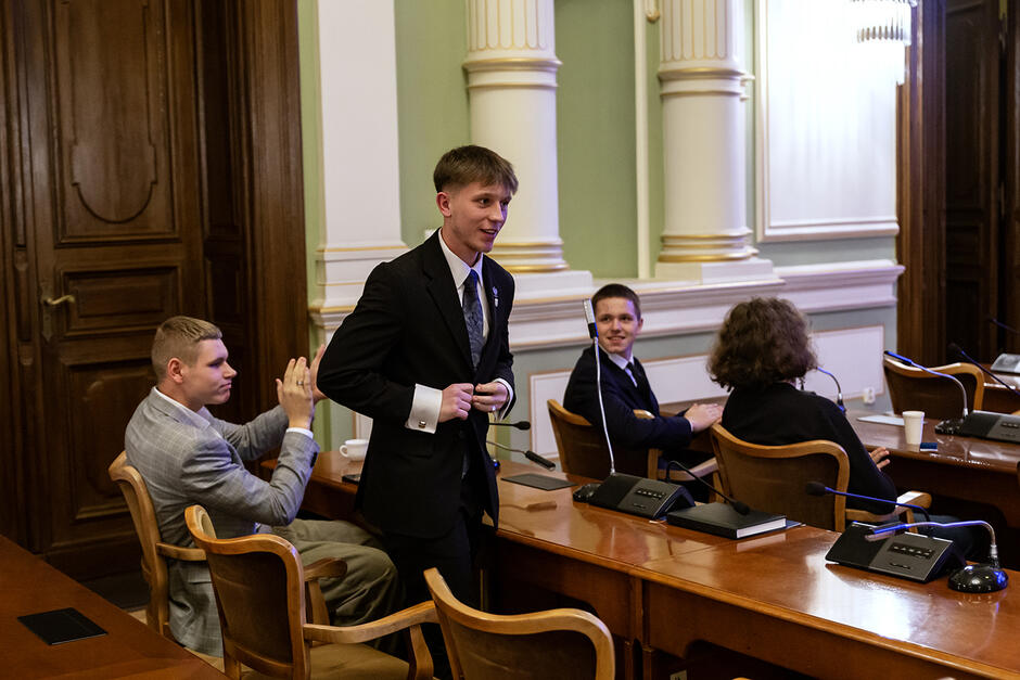 W sali obrad Młodzieżowej Rady Miasta Gdańska radni siedzą w ławach, a jeden z nich wstaje i się śmieje. Jeden z radnych bije mu brawo.