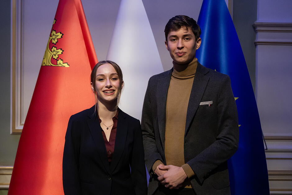 Dziewczyna i chłopak pozują do fotografii na tle flag w sali obrad Młodzieżowej rady Miasta Gdańska.