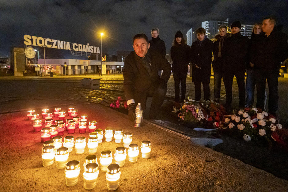 Noc, Na placu świecą się znicze, tuż za nimi kuca mężczyzna, trzyma w dłoni świecący znicz. Po prawej za nim stoi kilkanaście osób. W tle brama z napisem: STOCZNIA GDAŃSKA