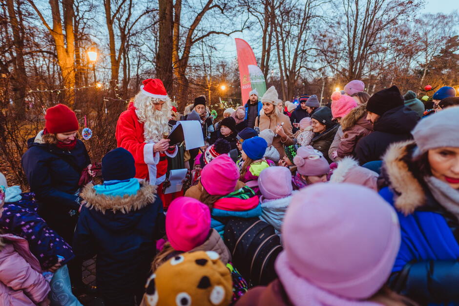 Na zdjęciu Święty Mikołaj rozdaje prezenty i czyta listy w otoczeniu dużej grupy dzieci i dorosłych, co tworzy świąteczną atmosferę. W tle widać zimowy park, udekorowany lampkami i świątecznymi ozdobami, a uczestnicy są ciepło ubrani w zimowe kurtki i czapki.