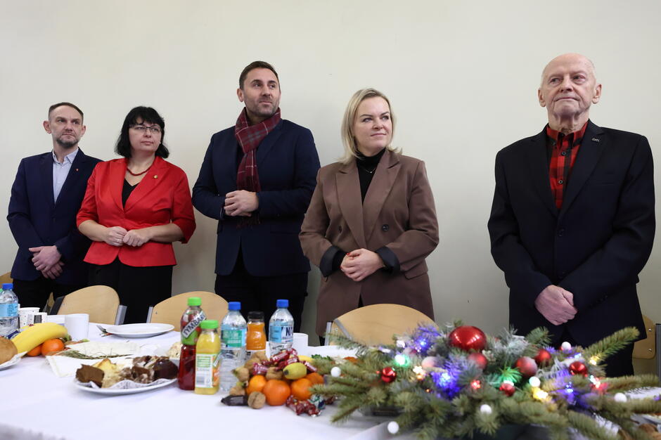 Na zdjęciu widzimy grupę osób stojących przy świątecznie udekorowanym stole, na którym znajdują się owoce, ciasta i napoje. Osoby są elegancko ubrane i wyglądają na uczestników oficjalnego spotkania, prawdopodobnie związanego z okresem świątecznym.