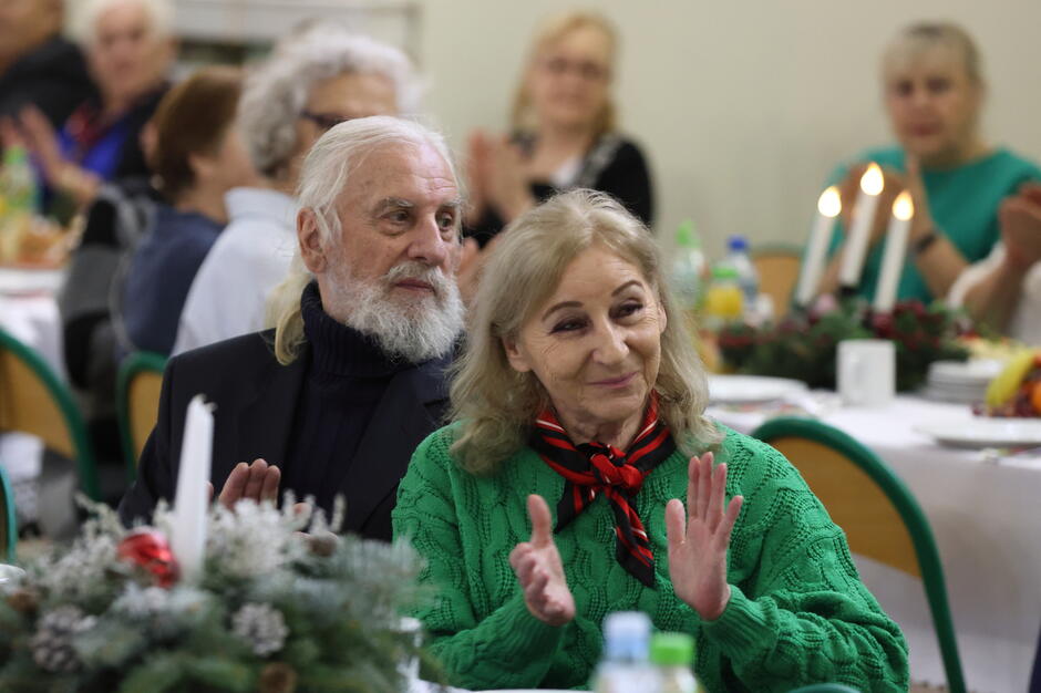 Na zdjęciu znajduje się para starszych osób siedzących przy świątecznie udekorowanym stole. Kobieta w zielonym swetrze i czerwono-czarnym szaliku oraz mężczyzna z siwą brodą i długimi włosami wyglądają na zadowolonych, uczestnicząc w uroczystym spotkaniu.