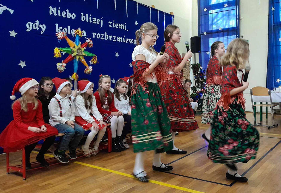 Na zdjęciu widać dzieci w strojach świątecznych i ludowych występujące podczas uroczystości, prawdopodobnie w związku z okresem bożonarodzeniowym. W tle znajduje się dekoracja z napisem „Niebo bliżej ziemi, gdy kolęda rozbrzmiewa” oraz kolorowa gwiazda, co podkreśla tradycyjny charakter wydarzenia.