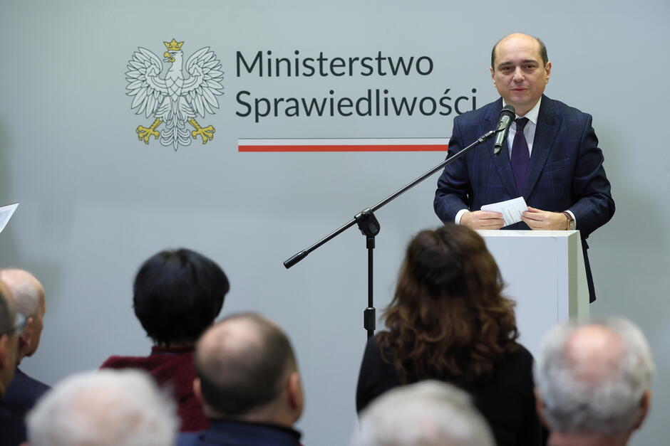 Na zdjęciu widzimy mężczyznę przemawiającego przy mównicy na tle logotypu Ministerstwo Sprawiedliwości  z godłem Polski. W tle znajduje się publiczność, która słucha wystąpienia