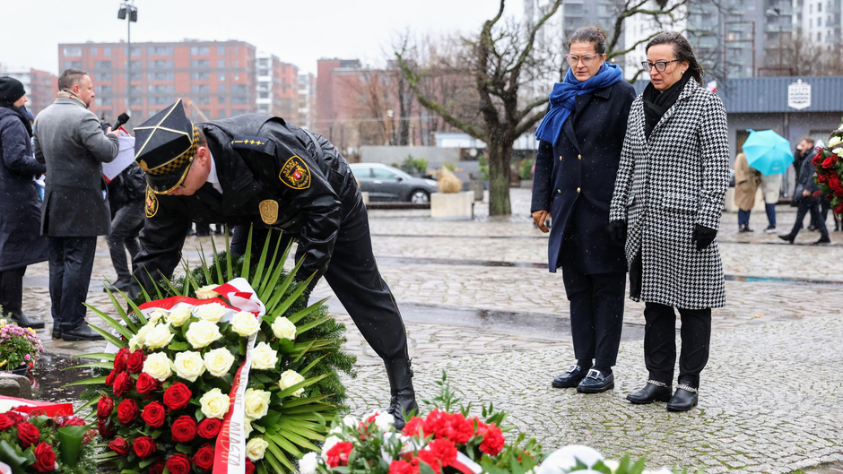 Zdjęcie przedstawia scenę upamiętniającą, która odbywa się na zewnątrz w deszczowy dzień. W centrum kadru widoczny jest funkcjonariusz w mundurze z charakterystycznym czarno-żółtym nakryciem głowy, układający duży wieniec z białych i czerwonych róż oraz zielonych liści na brukowanej powierzchni. Wieniec ma biało-czerwoną wstęgę z napisem. Za nim stoją dwie osoby, najprawdopodobniej oficjalni goście. Kobieta po lewej stronie ma na sobie ciemny płaszcz i niebieski szal owinięty wokół szyi, a także okulary. Druga kobieta, po prawej stronie, ubrana jest w płaszcz w biało-czarną pepitkę, czarne rękawiczki i okulary. Obie kobiety mają poważny i skupiony wyraz twarzy.  W tle widać kilka innych osób, część z nich trzyma parasole, a także miejskie budynki, drzewa i zaparkowane samochody. Sceneria sugeruje, że jest to uroczystość upamiętniająca, być może związana z hołdem dla poległych lub ważnym wydarzeniem historycznym. Atmosfera jest refleksyjna i formalna.