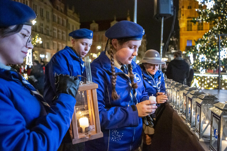 Zdjęcie przedstawia grupę harcerzy w niebieskich mundurach i beretach, którzy biorą udział w uroczystym przekazywaniu Betlejemskiego Światła Pokoju. Harcerze stoją wokół stołu z latarniami i świecami, trzymając płomień, podczas gdy w tle widać oświetlone choinki i zabytkowe budynki, co nadaje scenie świątecznego charakteru.