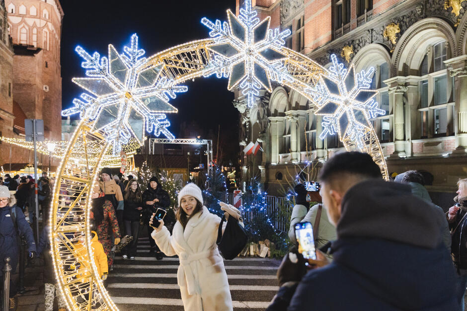 Zdjęcie przedstawia scenę z jarmarku bożonarodzeniowego, gdzie główną atrakcją jest ozdobny łuk z gigantycznymi podświetlonymi śnieżynkami. Na pierwszym planie uśmiechnięta kobieta w białym płaszczu pozuje do zdjęcia, podczas gdy tłum ludzi i świąteczne dekoracje tworzą tło pełne radosnej atmosfery