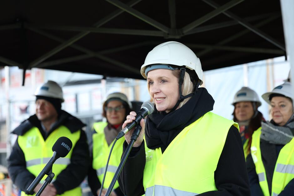 na zdjęciu kobieta w jasnych włosach, w białym kasku budowlanym na głowie, mówi do mikrofonu