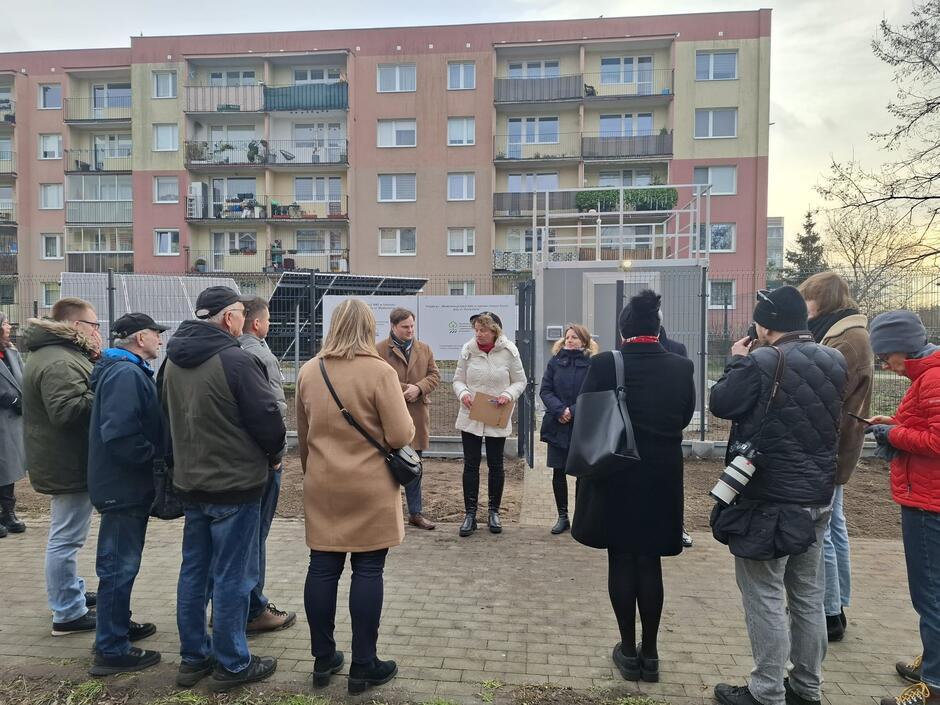 Na zdjęciu widzimy grupę osób stojących na chodniku przed budynkiem mieszkalnym, uczestniczących w wydarzeniu, które prawdopodobnie dotyczy projektu lub inwestycji. Na pierwszym planie widać osoby w ciepłych ubraniach, a jedna z nich trzyma aparat fotograficzny, co sugeruje dokumentację medialną.