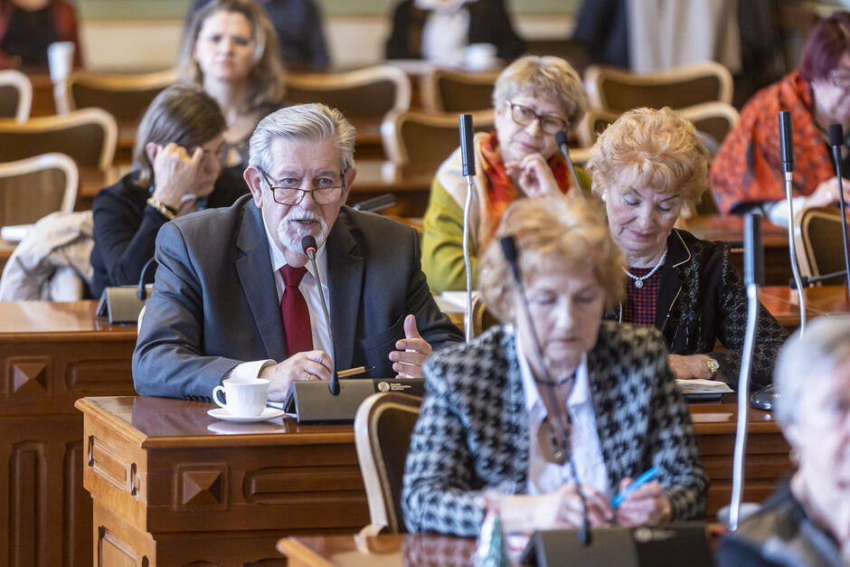 na zdjęciu kilku seniorów, siedzą za dużymi drewnianymi brązowymi stołami, widać starszego mężczyznę mówiącego do mikrofonu