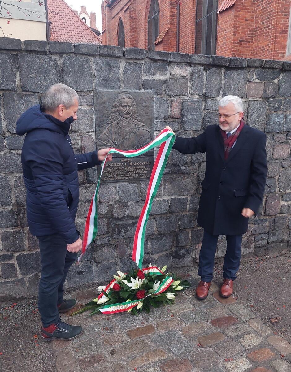 Dwaj mężczyźni po dwóch stronach tablicy, po lewej i prawej. Wokół tablicy są wstęgi w barwach narodowych Węgier 