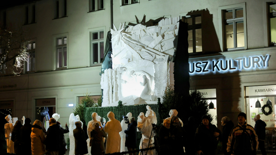 Na zdjęciu przedstawiona jest scena nocna w mieście, gdzie grupa ludzi zgromadziła się wokół dużej, podświetlonej rzeźby wykonanej prawdopodobnie z lodu lub śniegu. Rzeźba ma skomplikowaną strukturę z geometrycznymi wzorami oraz centralnym motywem przedstawiającym postacie ludzkie. Ludzie wokół rzeźby są ubrani w ciepłe zimowe ubrania, co sugeruje, że wydarzenie ma miejsce zimą. W tle widać budynek z napisem „RATUSZ KULTURY”, co może wskazywać na ośrodek kultury lub miejsce związane z wydarzeniami artystycznymi. Obok znajduje się kawiarnia z dużymi oknami udekorowanymi świątecznymi ozdobami, co sugeruje okres okołoświąteczny.  Niektórzy uczestnicy robią zdjęcia lub filmują rzeźbę, a inni przyglądają się jej z bliska. Sceneria jest dodatkowo oświetlona lampkami, tworząc świąteczną i uroczystą atmosferę.