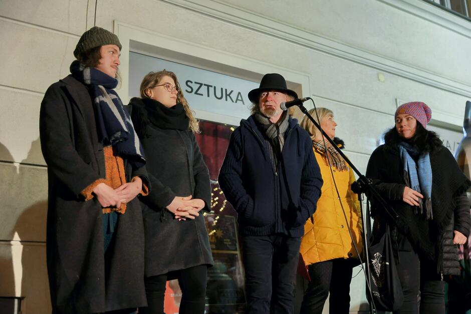 Na zdjęciu widzimy grupę pięciu osób stojących na scenie przed mikrofonem podczas wydarzenia o charakterze artystycznym lub kulturalnym. W tle znajduje się napis SZTUKA  (sztuka), co wskazuje na kontekst związany ze sztuką lub kulturą. Dekoracje świetlne i plakaty sugerują uroczysty charakter wydarzenia, być może w okresie świątecznym lub zimowym.