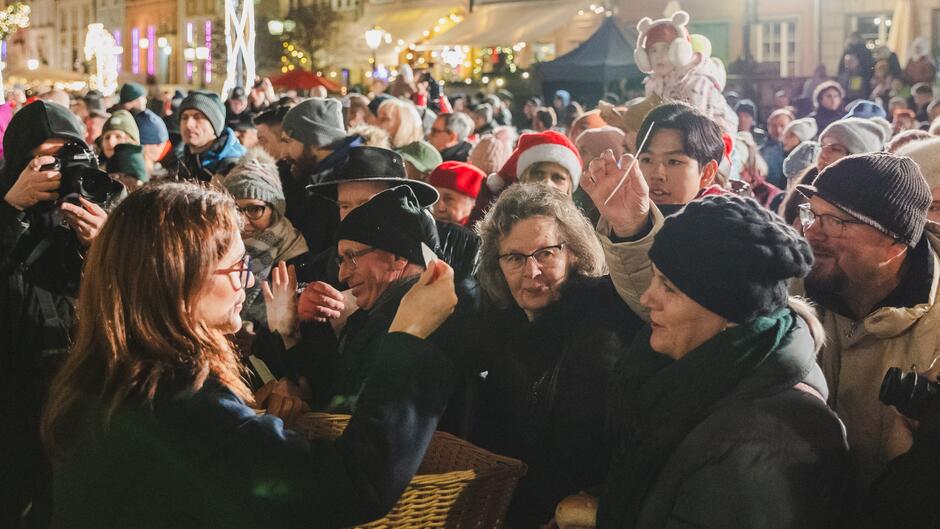 Na zdjęciu widzimy dużą grupę ludzi zgromadzonych na jarmarku świątecznym lub innym wydarzeniu odbywającym się na świeżym powietrzu w okresie zimowym. W tle można dostrzec udekorowane światłami budynki oraz stragany, co dodaje świątecznego klimatu. Na pierwszym planie kobieta w okularach i ciemnym płaszczu rozdaje coś z koszyka, a wokół niej zebrał się tłum ludzi, którzy wyciągają ręce, aby coś otrzymać. W tłumie można zauważyć osoby w zimowych ubraniach, takich jak czapki, szaliki i kurtki. Niektórzy mają na głowach świąteczne czapki Mikołaja.  Z tyłu widać również osoby robiące zdjęcia lub nagrywające kamerami, a także dzieci, w tym jedno siedzące na ramionach dorosłego i ubrane w różową kurtkę oraz nauszniki. Cała scena emanuje atmosferą świątecznej radości i wspólnoty.