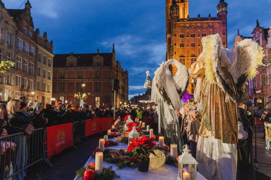 Na zdjęciu widzimy świąteczny jarmark lub uroczystość bożonarodzeniową w mieście, prawdopodobnie w Gdańsku, co sugerują czerwone banery z herbem i nazwą miasta. Scena odbywa się na tle zabytkowych kamienic oraz pięknie oświetlonej wieży ratusza. W centrum znajduje się udekorowany stół z lampionami, świecami, gałązkami świerku, czerwonymi poinsecjami oraz owocami, co tworzy świąteczną atmosferę. Obok stołu stoją osoby ubrane w stroje aniołów z dużymi skrzydłami i bogato zdobionymi szatami.  Zebrany tłum ludzi, w tym wielu z telefonami, fotografuje lub nagrywa wydarzenie. W tle widać również inne osoby przebrane w świąteczne kostiumy, co wskazuje na zorganizowane przedstawienie lub orszak. Cała scena jest rozświetlona światłami ulicznymi i dekoracyjnymi, podkreślając magiczny klimat wieczoru.