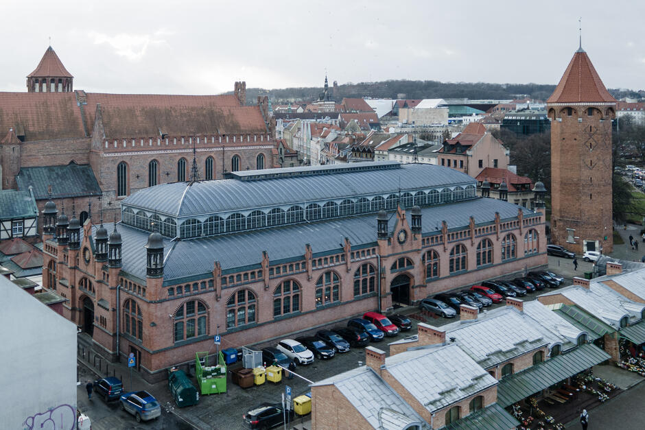 Zdjęcie przedstawia widok z lotu ptaka na zabytkową Halę Targową, która znajduje się w otoczeniu historycznej architektury. Budynek wykonany jest z czerwonej cegły i posiada duże, łukowate okna oraz metalowy dach z przeszklonymi elementami, co nadaje mu industrialno-gotycki charakter. Charakterystyczne wieżyczki i ozdobne detale architektoniczne podkreślają jego historyczny styl, prawdopodobnie z końca XIX lub początku XX wieku. W bezpośrednim sąsiedztwie hali znajduje się średniowieczny kościół z cegły o wysokim dachu i gotyckich oknach, a także ceglana wieża, która może być częścią dawnych fortyfikacji lub zabytkowym elementem obronnym.