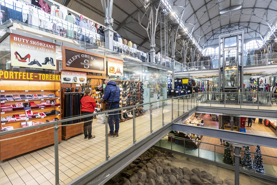 Zdjęcie przedstawia wnętrze Hali Targowej, która została zaadaptowana na nowoczesne centrum handlowe. Widoczna jest przestrzeń z antresolą, sklepami oraz szklaną windą umożliwiającą dostęp do różnych poziomów. W tle można zauważyć liczne stoiska z odzieżą, akcesoriami oraz innymi artykułami. Na pierwszym planie znajduje się sklep z galanterią skórzaną oferujący buty skórzane, paski, portfele oraz torebki. Drewniane półki wypełnione produktami nadają ciepły i elegancki wygląd, przyciągając klientów. Dwie osoby oglądają produkty, co wskazuje na aktywność handlową.