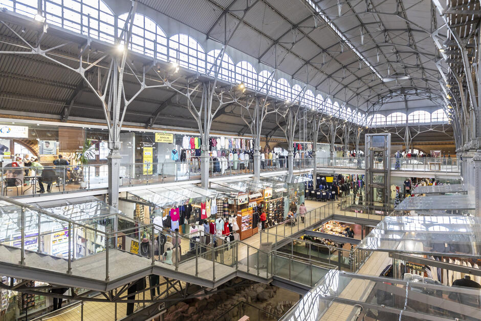 Zdjęcie przedstawia wnętrze Hali Targowej, która została przekształcona w nowoczesne centrum handlowe, łącząc zabytkową architekturę z funkcjonalnością współczesnych przestrzeni handlowych. Charakterystyczne cechy tego miejsca to wysokie, przeszklone łuki okien oraz ozdobne, metalowe filary i konstrukcje dachowe, które nadają wnętrzu industrialny, historyczny charakter. Hala składa się z kilku poziomów, z antresolą pełną sklepów odzieżowych, obuwniczych i akcesoryjnych. Na parterze znajdują się kolejne punkty sprzedaży oraz stoiska z żywnością i produktami regionalnymi. Widoczna jest także przeszklona winda oraz balustrady z przezroczystego szkła, które podkreślają nowoczesny wystrój wnętrza.