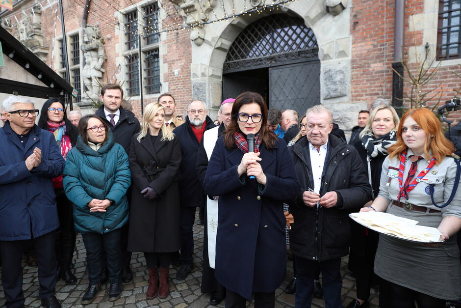 Na zdjęciu widzimy grupę osób zgromadzonych przed zabytkowym budynkiem z cegły, ozdobionym rzeźbami i świątecznymi lampkami. Na pierwszym planie znajduje się kobieta w ciemnym płaszczu i szaliku w kratkę, trzymająca mikrofon i przemawiająca do zebranych. Obok niej stoi starszy mężczyzna w czarnej kurtce, a po prawej stronie młoda kobieta w harcerskim mundurze trzymająca tacę z opłatkami. W tle znajduje się więcej osób w zimowych ubraniach, w tym mężczyźni i kobiety, niektórzy z nich mają na sobie eleganckie płaszcze. Atmosfera sugeruje uroczysty charakter wydarzenia, prawdopodobnie związany ze świętami Bożego Narodzenia, co podkreśla obecność opłatków.  Cała scena odbywa się na brukowanym dziedzińcu lub placu, a historyczna architektura budynku wskazuje, że może to być ważne miejsce publiczne lub kulturalne.