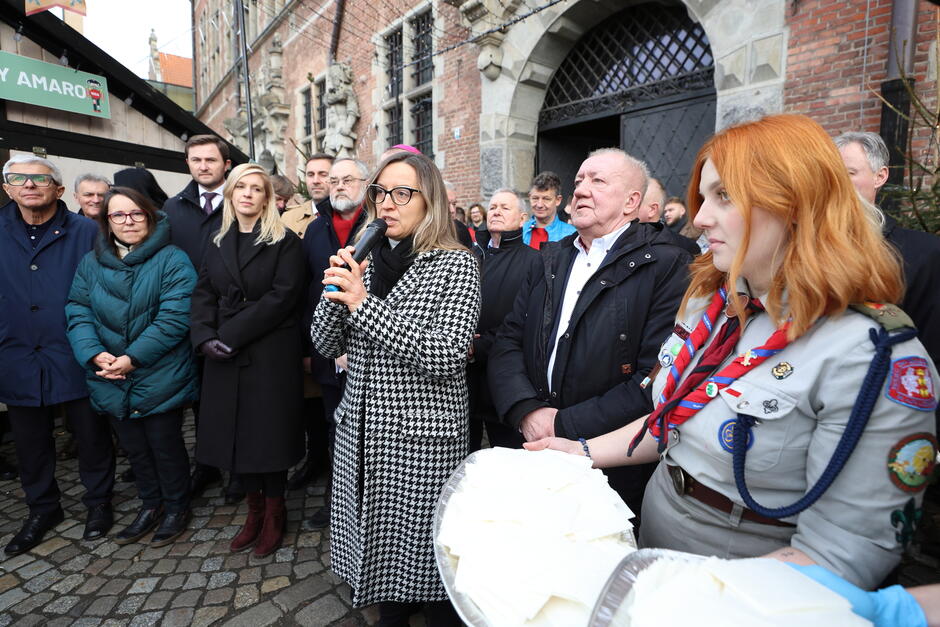 Na zdjęciu widzimy uroczystość odbywającą się na zewnątrz, przed zabytkowym budynkiem z cegły, ozdobionym rzeźbami. Na pierwszym planie kobieta w płaszczu w czarno-białą kratkę przemawia do zgromadzonych, trzymając mikrofon. Obok niej stoi młoda kobieta w harcerskim mundurze z odznakami i chustą, trzymająca tacę wypełnioną opłatkami, co sugeruje, że wydarzenie może być związane z tradycyjnym spotkaniem opłatkowym. Za nimi znajduje się grupa elegancko ubranych ludzi, w tym mężczyźni i kobiety w płaszczach i szalikach, co wskazuje na zimowy okres. W tle widoczna jest również drewniana budka, co może sugerować, że wydarzenie ma miejsce na jarmarku bożonarodzeniowym lub innym świątecznym spotkaniu. Atmosfera wydaje się formalna, ale jednocześnie ciepła i świąteczna.