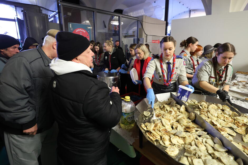 Na zdjęciu widzimy scenę z akcji charytatywnej lub świątecznego wydarzenia, gdzie harcerki w mundurach serwują jedzenie. Są one ubrane w szare koszule z odznakami, czerwone chusty i mają na rękach jednorazowe rękawiczki. Podają one pierogi z dużych, metalowych tac. Jedna z harcerek nakłada pierogi na talerze, a druga posypuje je przyprawami. Po lewej stronie znajdują się osoby czekające w kolejce na posiłek. Są one ciepło ubrane, co sugeruje zimową porę roku. W tle widoczne są dodatkowe osoby pomagające przy wydawaniu jedzenia oraz duży garnek, prawdopodobnie z zupą lub innym ciepłym daniem.  Atmosfera wydaje się przyjazna i wspólnotowa. Całość sugeruje świąteczne spotkanie z posiłkiem dla potrzebujących lub lokalnej społeczności, podkreślając troskę o innych w okresie Bożego Narodzenia.