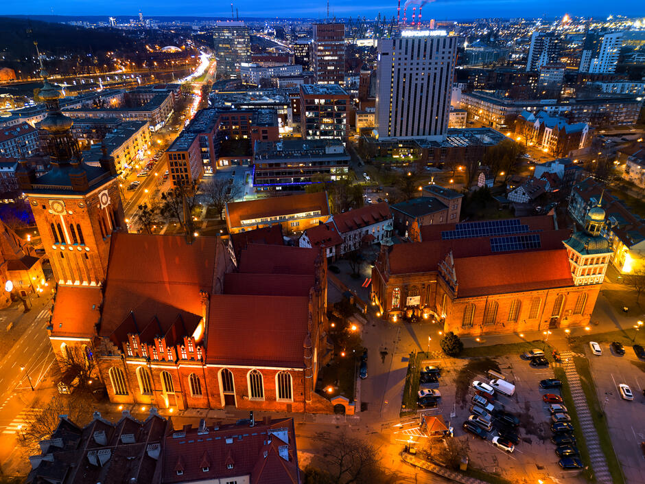 To zdjęcie przedstawia panoramę miasta o zmierzchu, ukazując historyczne budynki z czerwonymi dachami i wysokimi wieżami otoczone nowoczesnymi wieżowcami i ulicami rozświetlonymi latarniami. W centralnej części obrazu dominuje duży kościół z cegły, którego architektura kontrastuje z nowoczesną zabudową w tle