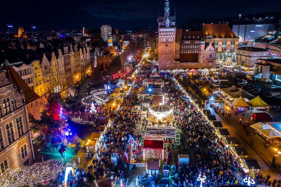 Widok z góry wieczorem: na dużym placu rozstawione budki, karuzela, wszystko oświetlone iluminacjami świątecznymi. Po lewej i w tle budynki