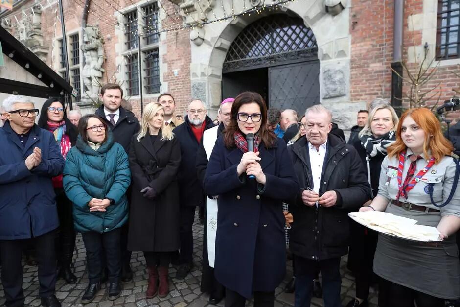 Kilkanaście osób stoi przed budynkiem, w środku kobieta w okularach z mikrofonem