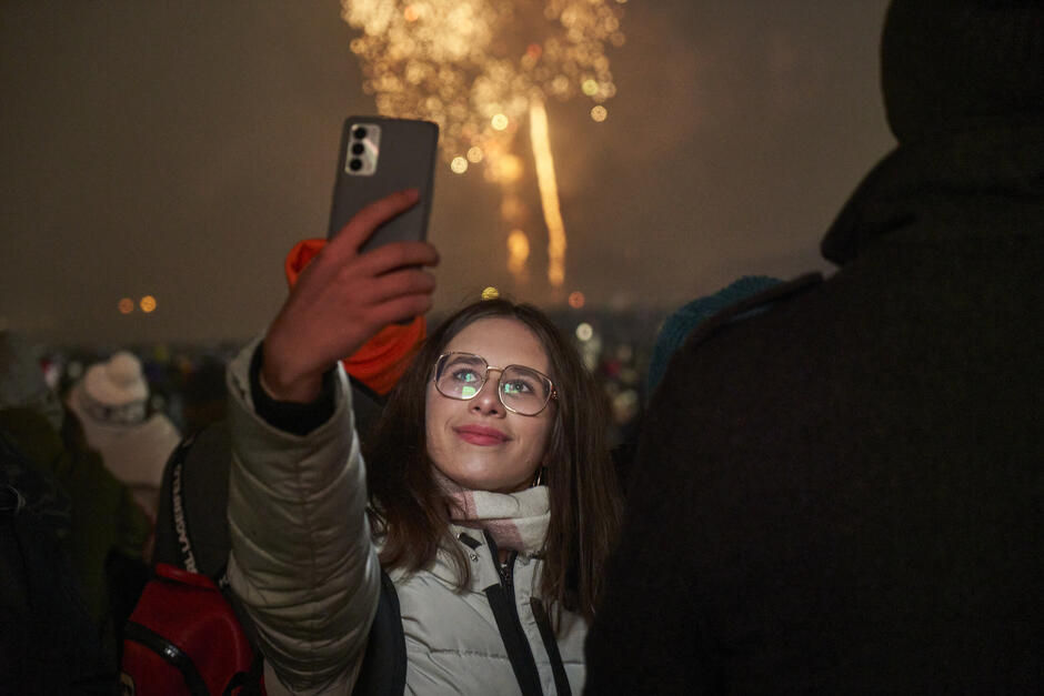 Na zdjęciu widać młodą kobietę robiącą selfie na tle nocnych fajerwerków. Jest uśmiechnięta, ubrana w ciepłą kurtkę i szalik, a wokół niej znajduje się tłum ludzi, co sugeruje, że uczestniczy w sylwestrowej imprezie plenerowej.