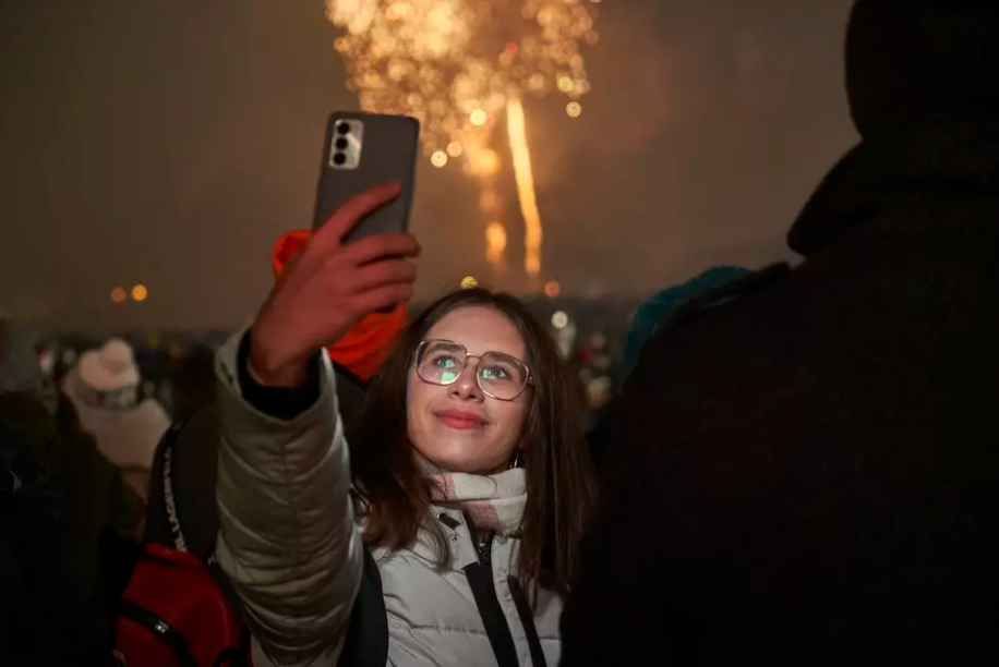 Zdjęcie przedstawia młodą kobietę robiącą selfie telefonem podczas pokazu fajerwerków. Kobieta nosi okulary, kurtkę puchową i szalik, a jej twarz jest oświetlona światłem z ekranu telefonu. W tle widoczny jest nocny krajobraz, na którym dominują jasne eksplozje fajerwerków na niebie oraz sylwetki innych osób obserwujących pokaz. Scena emanuje radosną, świąteczną atmosferą, związaną z celebracją Nowego Roku.