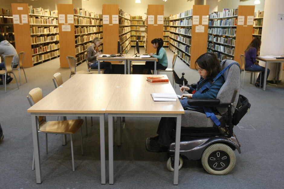 osoba na wózku w bibliotece, siedzi przy stole, wokół regały z książkami