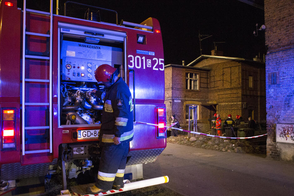 noc, wóz strażacki, strażak w kasku i kombinezonie na pierwszym planie, w tle budynek odgrodzony taśmą 