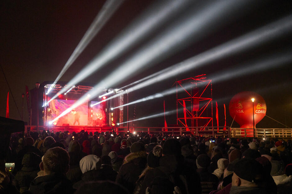 Ciemno, ludzie zebrani pod sceną na świeżym powietrzu. Ze sceny biją światła