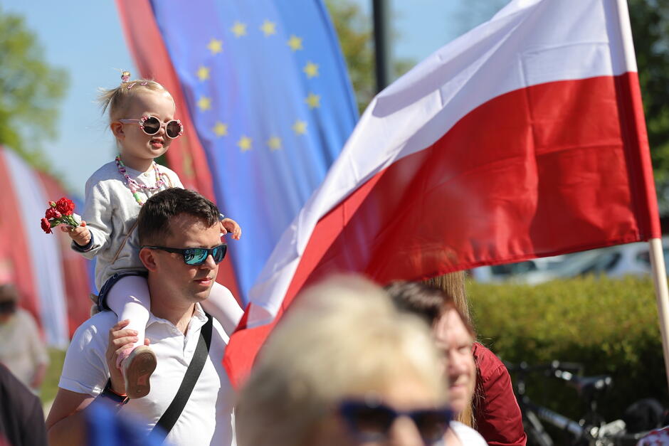 Na zdjęciu widzimy scenę z publicznego wydarzenia, prawdopodobnie obchodów lub manifestacji. Na pierwszym planie ojciec niesie na ramionach małą dziewczynkę, która trzyma w dłoni czerwone kwiaty. Dziewczynka ma na sobie okulary przeciwsłoneczne i naszyjnik, co dodaje jej uroku. W tle widoczne są flagi Polski (biało-czerwona) oraz Unii Europejskiej (niebieskie z żółtymi gwiazdami), co sugeruje, że wydarzenie ma związek z tematyką europejską lub polsko-unijną. Atmosfera jest pogodna, a zdjęcie wykonano w słoneczny dzień, co podkreślają jasne barwy i cienie.