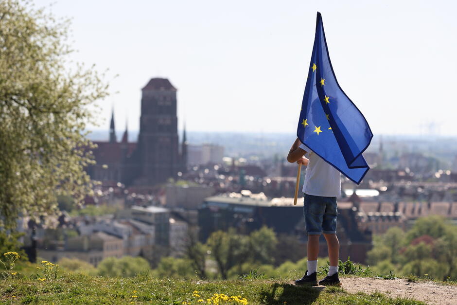 Na zdjęciu widzimy chłopca stojącego na wzgórzu, trzymającego dużą flagę Unii Europejskiej. Flaga powiewa na wietrze, a w tle rozciąga się panorama miasta z charakterystycznymi budynkami, w tym wysoką katedrą. Sceneria jest pełna zieleni, co wskazuje na wiosenną porę roku. Zdjęcie wykonane w słoneczny dzień, co podkreślają jasne barwy i wyraźne kontury. Chłopiec ubrany jest w letni strój: białą koszulkę i jeansowe szorty, co dodaje kompozycji swobody i radosnej atmosfery. Całość obrazu emanuje spokojem i nawiązuje do idei jedności oraz współpracy europejskiej.