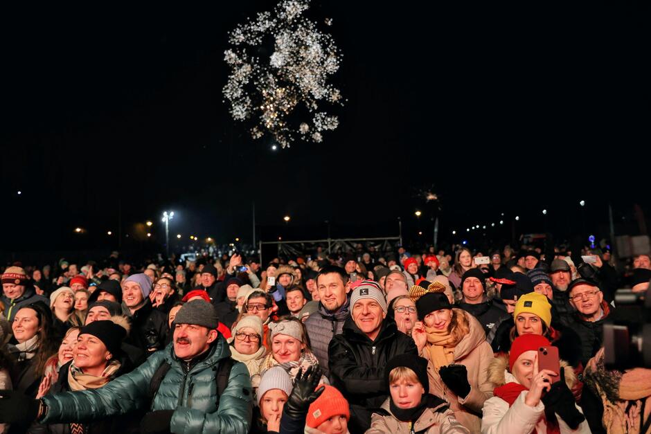 Na zdjęciu widać tłum ludzi zgromadzonych na wydarzeniu plenerowym nocą, wielu z nich ubranych jest ciepło, co sugeruje chłodną porę roku. Nad tłumem rozbłyska fajerwerk, a niektórzy uczestnicy robią zdjęcia lub nagrywają chwile telefonami.