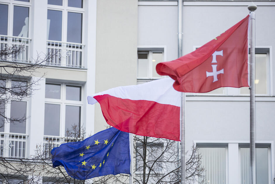 Na zdjęciu widoczne są trzy flagi powiewające na masztach przed budynkiem. Flaga Unii Europejskiej (na dole) - niebieska z dwunastoma żółtymi gwiazdami ułożonymi w okręgu. Flaga Polski (w środku) - biało-czerwona, symbol narodowy Polski. Czerwona flaga z białym krzyżem i dwoma mieczami (na górze) - prawdopodobnie jest to flaga miasta Gdańska lub innego lokalnego symbolu.