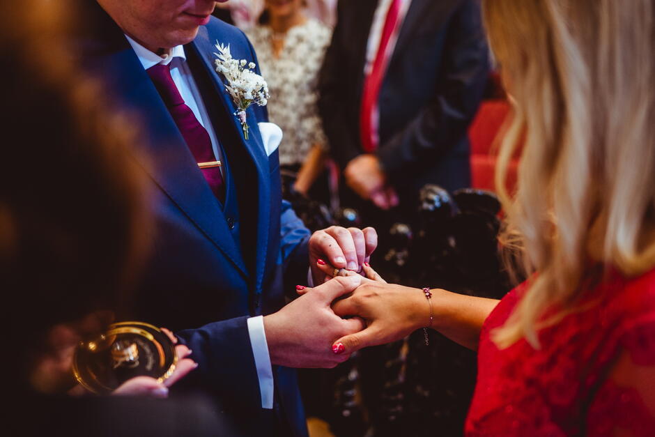 Na zdjęciu uwieczniono moment zakładania obrączki podczas ceremonii ślubnej. Mężczyzna w eleganckim granatowym garniturze wkłada obrączkę na palec kobiety ubranej w czerwoną suknię, co symbolizuje zawarcie małżeństwa.