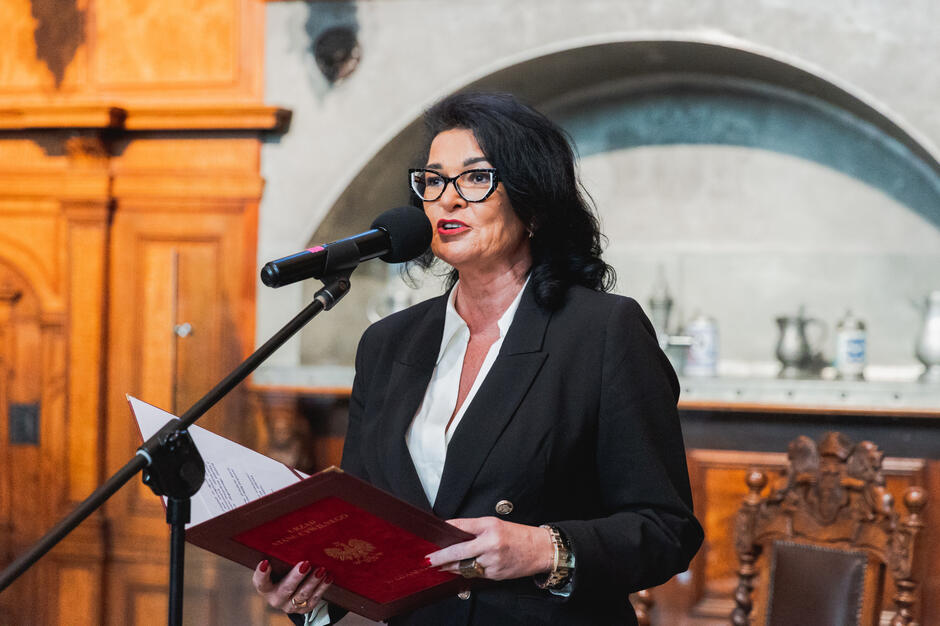 Na zdjęciu kobieta w eleganckim stroju stoi przy mikrofonie, trzymając w rękach dokument w czerwonej oprawie z godłem Polski, co sugeruje oficjalną uroczystość. Za nią widoczna jest stylowa, drewniana aranżacja wnętrza, nadająca poważny charakter wydarzeniu.