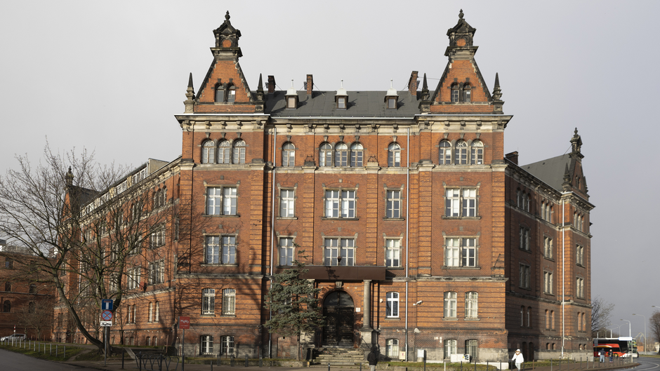 Na zdjęciu widoczny jest zabytkowy budynek z czerwonej cegły, o architekturze nawiązującej do stylu neogotyckiego lub historyzmu. Posiada on liczne ozdobne detale, takie jak wieżyczki, zdobione gzymsy oraz wysokie okna z łukami. Wejście główne znajduje się na środku fasady, prowadzą do niego schody, a nad drzwiami znajduje się niewielki daszek wsparty na kolumnach. Budynek sprawia wrażenie dawnej szkoły, urzędu lub innej instytucji publicznej.  Otoczenie obejmuje chodniki, drzewa oraz znaki drogowe, co wskazuje na miejskie położenie. W tle widoczny jest autobus, co sugeruje bliskość komunikacji publicznej. Całość ma reprezentacyjny charakter, co może świadczyć o historycznym znaczeniu tego obiektu.       