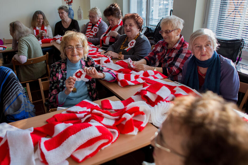 grupa seniorów dzierga szaliki na paradę niepodległości, na stolach leżą biało - czerwone szaliki