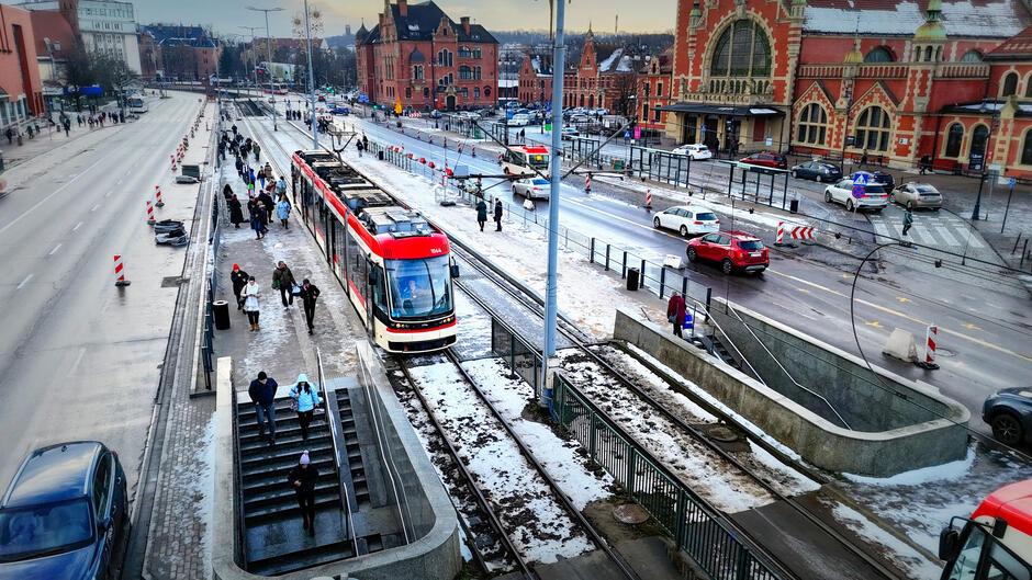 Zdjęcie przedstawia zimowy dzień w Gdańsku, ukazując ruchliwą ulicę z przystankiem tramwajowym oraz pieszymi korzystającymi z komunikacji miejskiej. W tle widoczny jest zabytkowy budynek Dworca Głównego, będący charakterystycznym elementem architektury miasta.