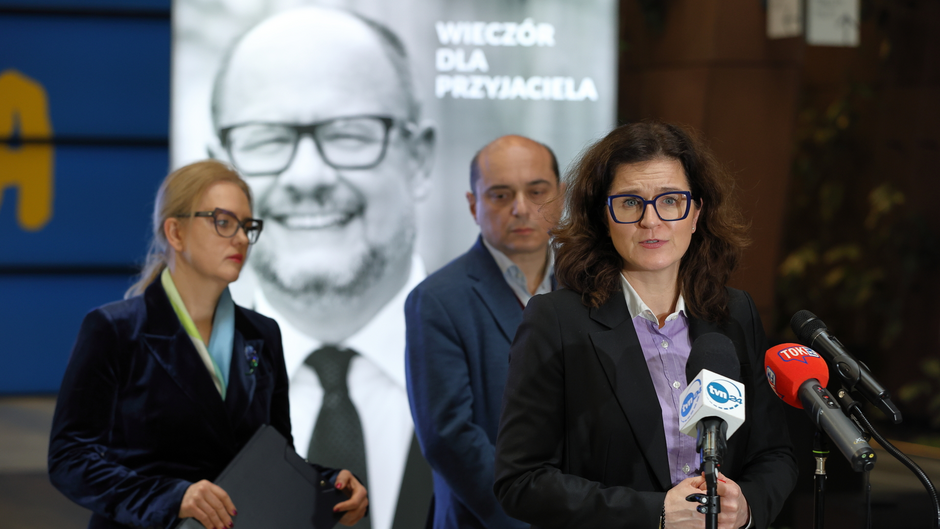 Na zdjęciu widzimy konferencję prasową lub briefing. W centrum znajduje się kobieta w okularach, ubrana w czarną marynarkę i jasnofioletową koszulę, która przemawia do mikrofonów oznaczonych logotypami mediów, takich jak TVN24 i TOK FM. Za nią stoją dwie inne osoby – kobieta w okularach, ciemnym stroju i apaszce oraz mężczyzna w marynarce. W tle widoczny jest duży czarno-biały portret uśmiechniętego mężczyzny w okularach i garniturze oraz napis w języku polskim „WIECZÓR DLA PRZYJACIELA” (co oznacza „Wieczór dla przyjaciela”). Scena sugeruje, że może to być wydarzenie poświęcone pamięci tej osoby, być może zmarłej lub uhonorowanej w szczególny sposób.  Całe wydarzenie wygląda na formalne, prawdopodobnie związane z polityką, kulturą lub działalnością społeczną.