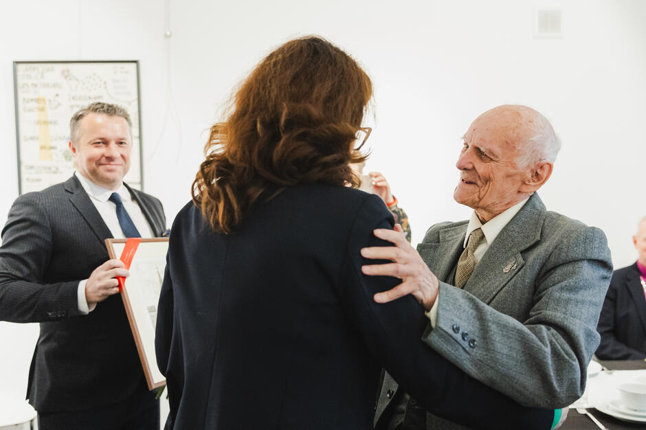 Na zdjęciu widać uroczyste spotkanie, podczas którego starszy mężczyzna w eleganckim garniturze wita się serdecznie z kobietą ubraną w ciemny strój. W tle znajduje się uśmiechnięty mężczyzna trzymający oprawiony dyplom, co sugeruje oficjalną ceremonię wręczenia wyróżnień lub podziękowań.